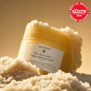 Start 54 Naturium The Glow Getter Multi Oil Body Scrub - odżywczy peeling cukrowo-olejkowy do ciała w jasnożółtym słoiczku, otoczony połyskującymi kryształkami cukru na ciepłym, złocisto-beżowym tle. W prawym górnym rogu czerwone logo nagrody Allure Best of Beauty 2026.