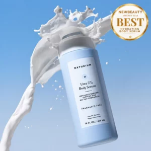 Start 53 Niebieska butelka serum do ciała Naturium Urea 5% Body Serum z 5% mocznikiem, otoczona efektownym rozbryzgiem białej, kremowej emulsji na błękitnym tle. W prawym górnym rogu złoty medal NewBeauty Awards 2026 za najlepsze nawilżające serum do ciała.